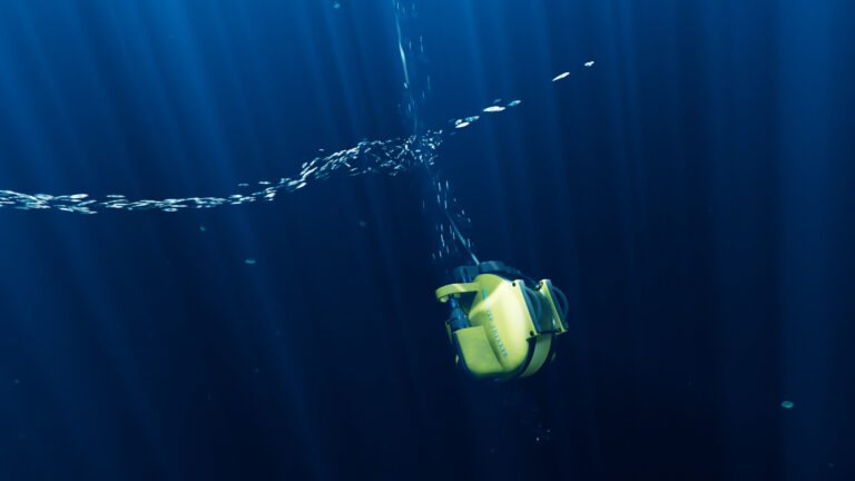 Deep Trekker: ROV DTG3: Gul undervandsdrone med Deep Trekker-logo hænger i et kabel i dybblåt vand med solstråler og en lang stime af sølvfisk